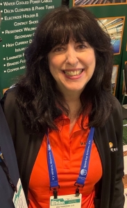 A woman with long, dark hair is smiling brightly at the camera. She is wearing an orange Watteredge polo shirt, a black jacket, and a conference badge on a blue lanyard. A promotional backdrop featuring industrial products is behind her.
