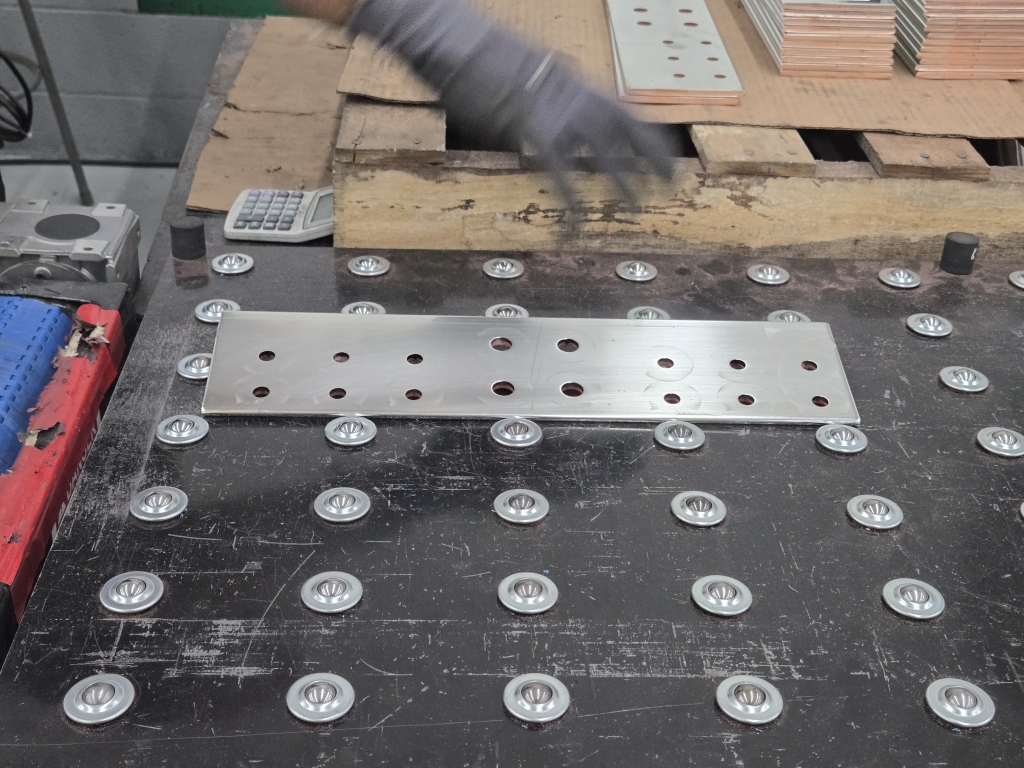 A rectangular tin-plated copper bus bar with evenly spaced holes lies on a perforated black metal surface. A gloved hand is visible in motion near the top, and additional metal components and wooden pallets are in the background.