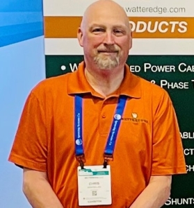 A bald man with a trimmed beard wearing an orange Watteredge polo shirt and a conference badge stands in front of a Watteredge promotional display. He is smiling slightly and looking at the camera.