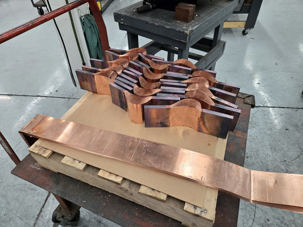 A stack of thick, curved copper shunts with darkened weld marks is arranged on a workbench in a manufacturing facility. A long flat copper piece extends outward from the table.