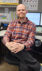 A bald man with a trimmed beard wearing a red and blue plaid shirt sits in an office chair with hands folded, smiling at the camera. Computer monitors, a phone, and office charts are visible in the background.