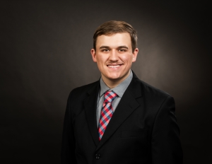 A young man with short brown hair is smiling in a professional studio portrait. He is wearing a black suit jacket, a gray shirt, and a red, white, and blue plaid tie, posed against a dark background.