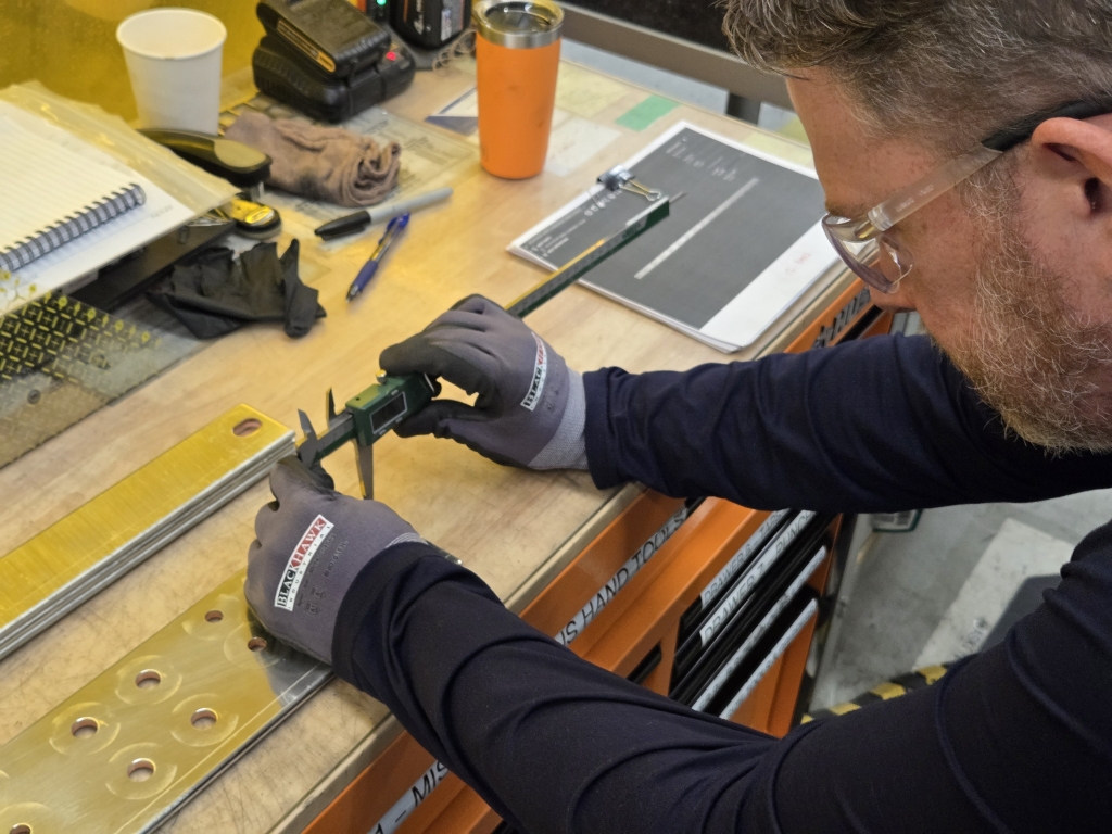 A Watteredge employee uses a digital caliper to measure a punched bus bar on a workbench, ensuring accuracy before production continues.