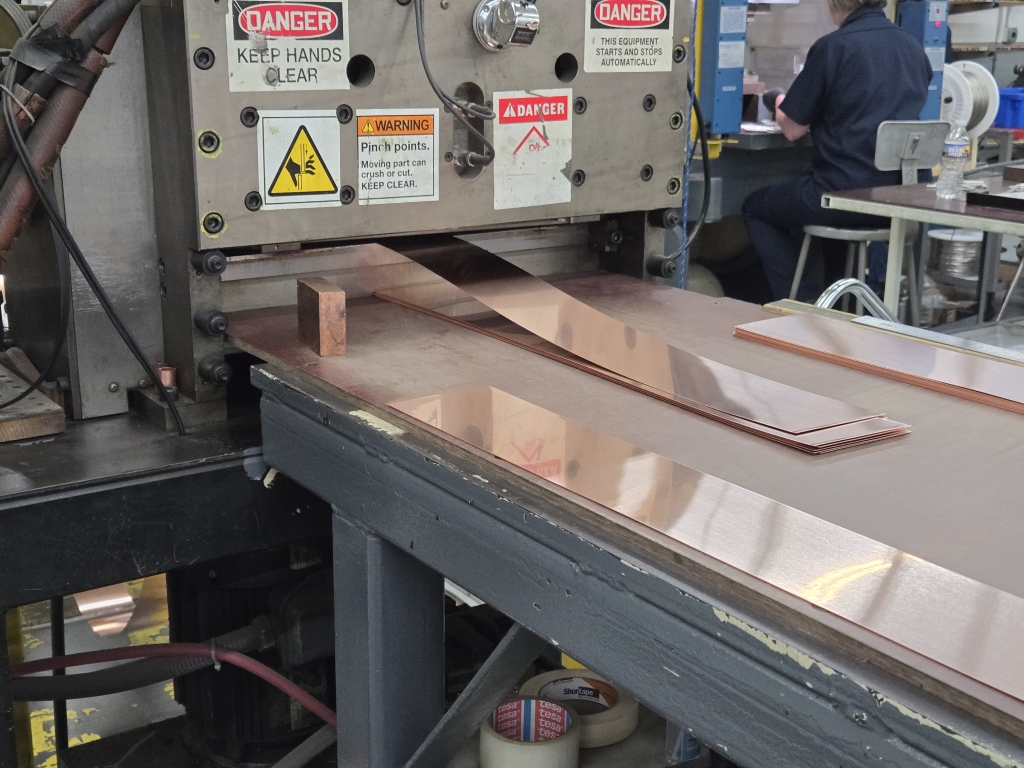 Copper sheets are being fed into a large industrial shear machine, with safety warning labels visible on the equipment. A worker is seated in the background at a separate workstation.