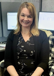 A woman with shoulder-length blonde hair wearing a black floral dress and a black cardigan is seated at an office desk, smiling at the camera. Two computer monitors are visible in the background.