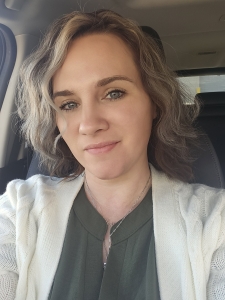 A woman with wavy, shoulder-length hair is sitting in a car, wearing a green top and a cream-colored cardigan. She is looking at the camera with a soft smile.