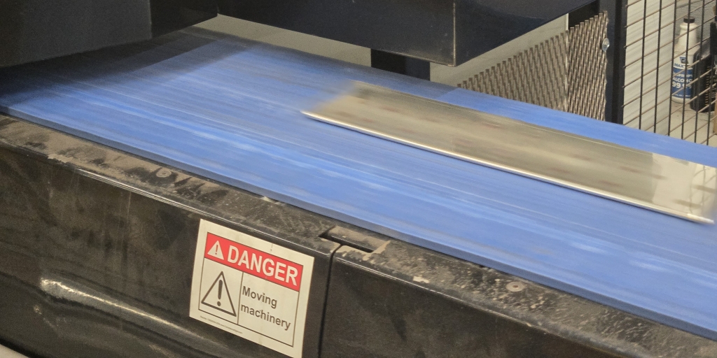 A tin-plated copper bus bar travels on a blue conveyor belt after processing in Watteredge's punching machine, with a visible warning label indicating moving machinery.
